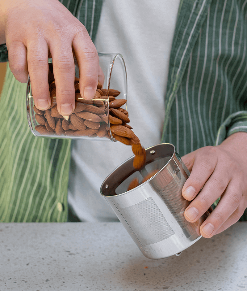 Person pouring almonds from a glass container into a stainless steel filter
